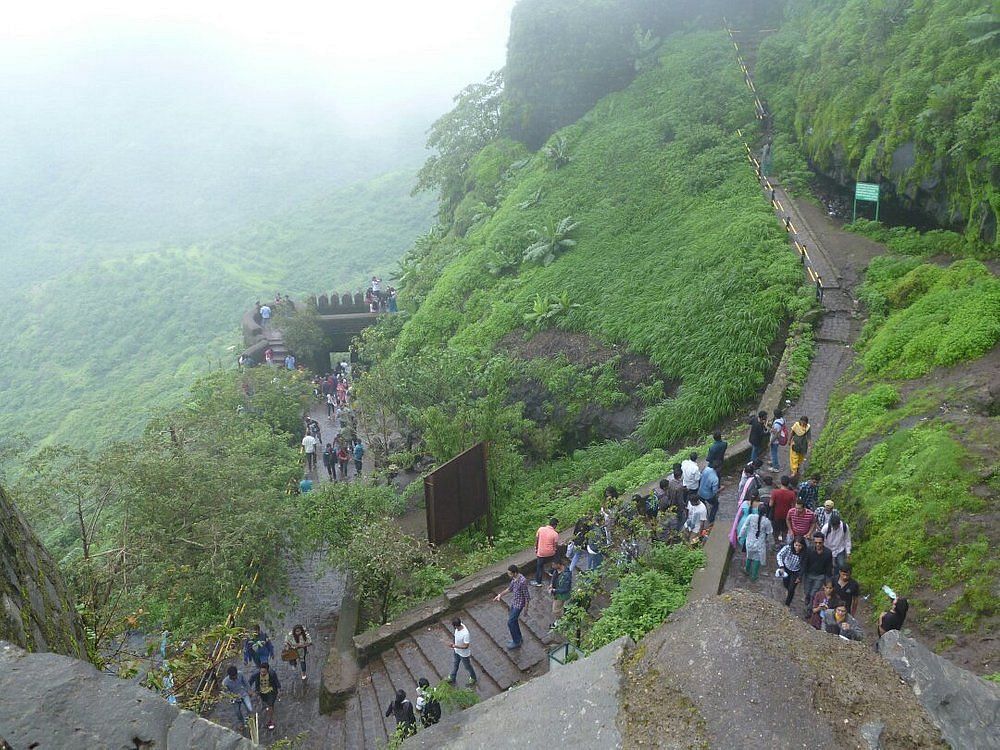 A winding, stone staircase climbs steeply up a lush, green hillside. People are walking up the stairs, and clouds are visible in the sky above.